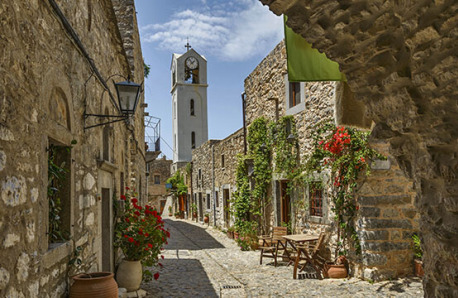 Cobbled street with traditional stone buildings with plants on the walls and a clock tower in the background. Sunshine.