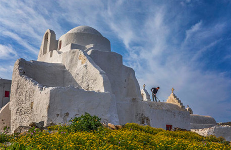 White traditional church with a dome surrounded by wildflowers. A man is painting. Sunny with light clouds.