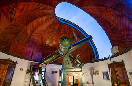 A man inside an observatory with a large telescope. The observatory has a beautiful wooden dome ceiling with an opening, allowing the telescope to point towards the sky.