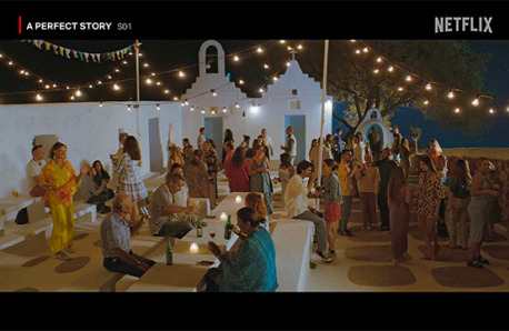 Scene from "A Perfect Story" Season 1 on Netflix showing a group of people at an outdoor evening party with festive lights, near a traditional white building.