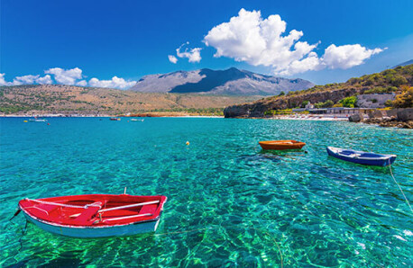 Sea with crystal clear waters and small boats. In the background beach with sunbeds and mountains.