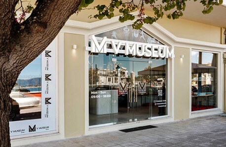 Γυάλινη πρόσοψη μουσείου με το όνομα του μουσείου, πλαισιωμένη από ανοιχτόχρωμο τοίχο. Αντανακλά χαμηλά κτίρια και καθαρό ουρανό.