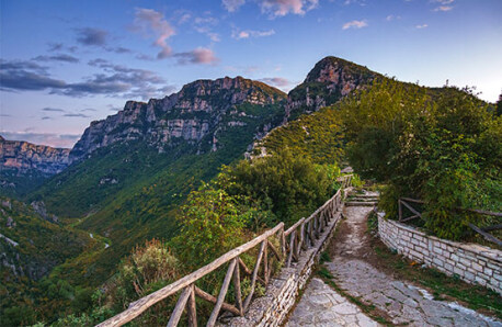 Paved path with a wooden fence and a view of the canyon. Green steep slopes and high cliffs. Clear sky with few clouds in the evening.