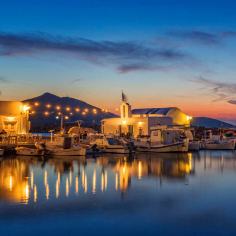 Peaceful harbor with boats and a small white church with lights at sunset. 