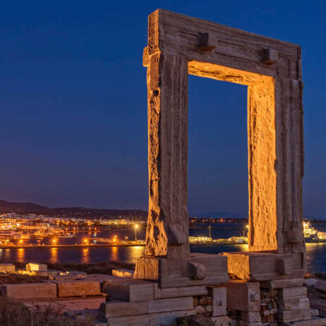 Night view of a large ancient gate in the foreground and in the background a Cycladic settlement with a port. 