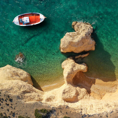 Aerial view of a white and red boat in crystal clear sea next to small beaches surrounded by rocks. 