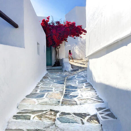 Girl walking on cobbled alley between white houses. In a yard a large red bougainvillea. 