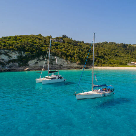 Beach of the island with pine trees overlooking the greenish blue waters and two sailing boats in the background