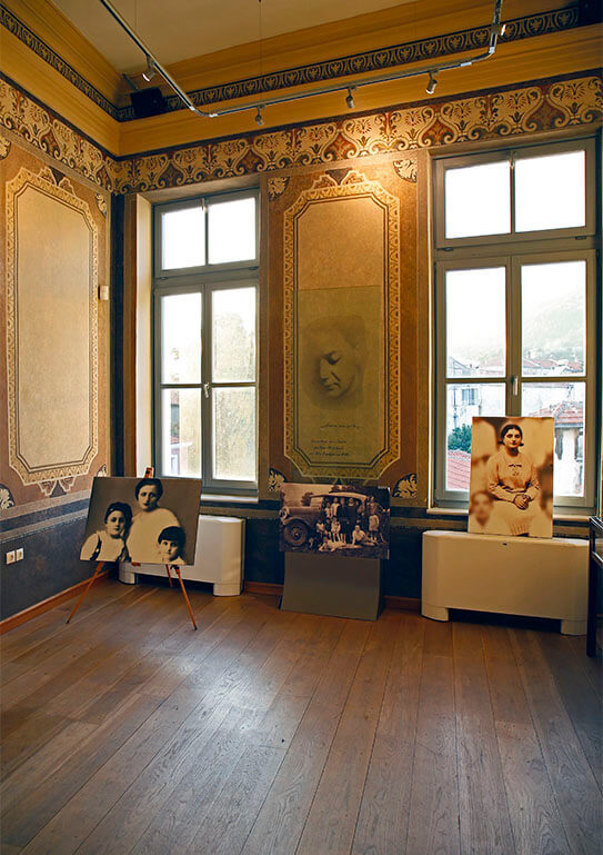 High ceiling hall with painted walls, large windows and wooden floor. Hatzidakis' picture on the wall and large family photos in front.