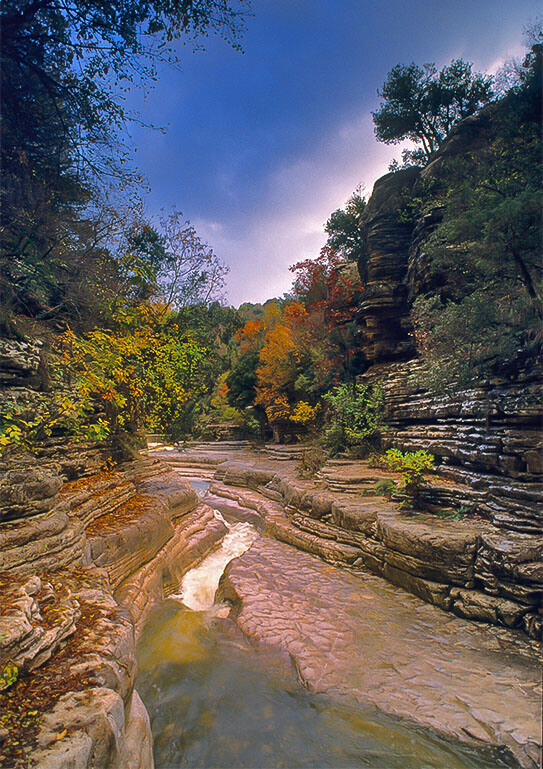Small river that runs between rocks that form lines. Dense vegetation all around. Blue sky with few clouds.