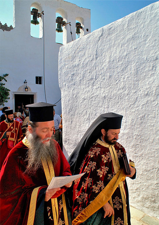 Priests in festive Orthodox attire emerge from a white church with bells and walk down an alley with a white wall. Sunshine.