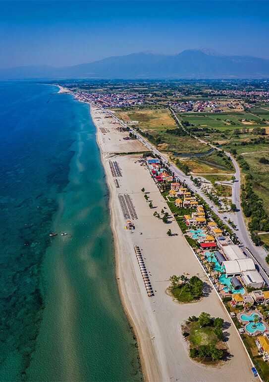 Aerial view of a long sandy beach with turquoise waters, lined with resorts and green fields stretching into the distance.