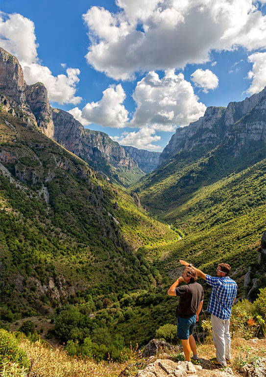 Two men observe the Vikos gorge from below. Green steep slopes and high cliffs. Blue sky with few clouds.