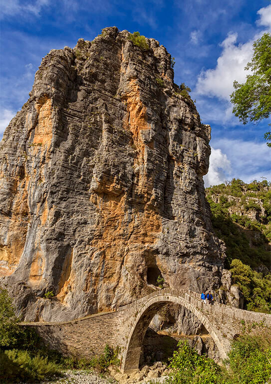 Stone arch bridge with people crossing, set against a towering rocky cliff under a blue sky.