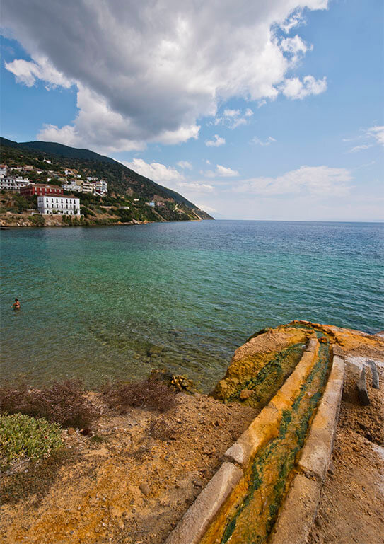 Seaside landscape with yellowish rocks and a stream that ends in the sea. In the background, a wooded slope with a settlement. Summer day with few clouds.