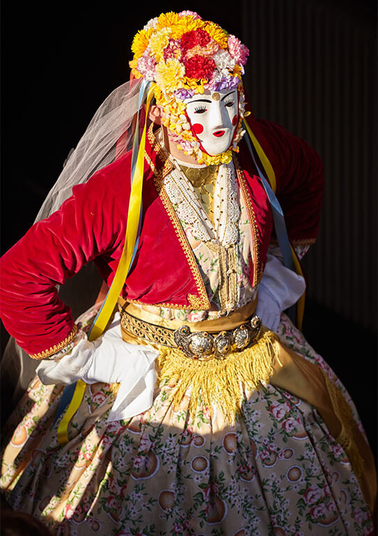 Man in traditional Boula carnival costume. With a red jacket, white female mask, flowers on his head and a veil.