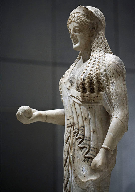 Ancient marble female statue with ornate hair and folds in her clothing, in a museum space with special lighting.