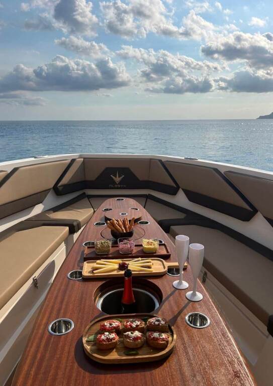 Elegant dining setup on the bow of a yacht with a charcuterie board, cupcakes, and champagne flutes; ocean horizon in the background under a clear blue sky.