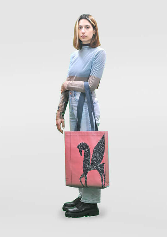 Person standing with a tote bag featuring a black sequined seahorse design, wearing a light blue top, gray trousers, and black platform shoes