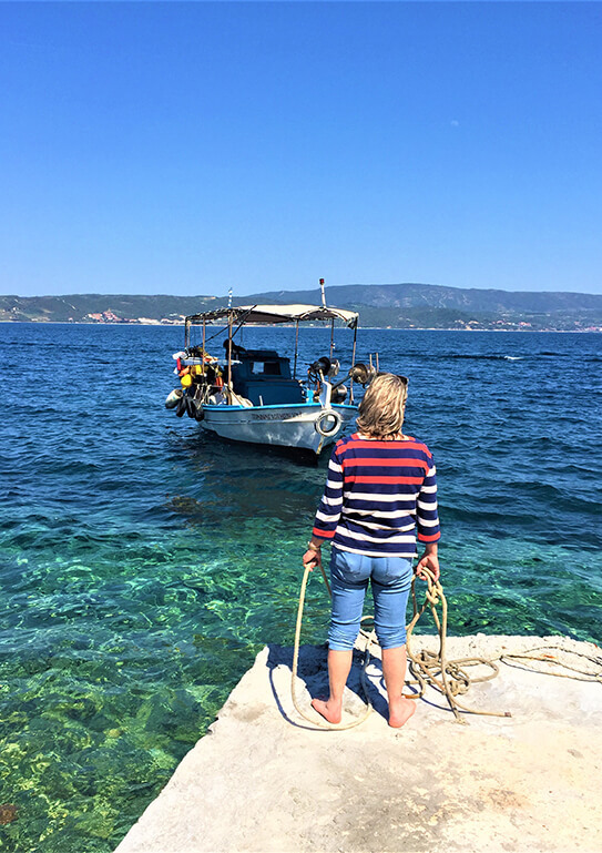 Person standing on a rocky shore holding a rope, watching a boat on clear blue water with hills in the distance.