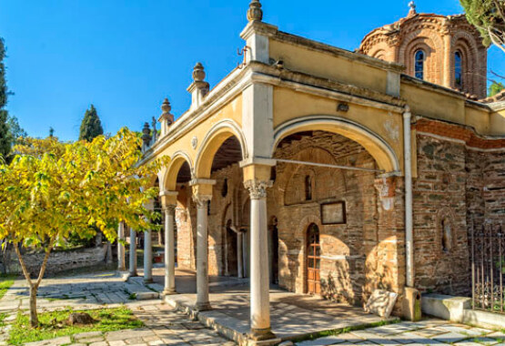 Βυζαντινή πέτρινη εκκλησία με τρούλο και καμάρες στο προαύλιο. Πλακόστρωτη αυλή με μικρό δέντρο.