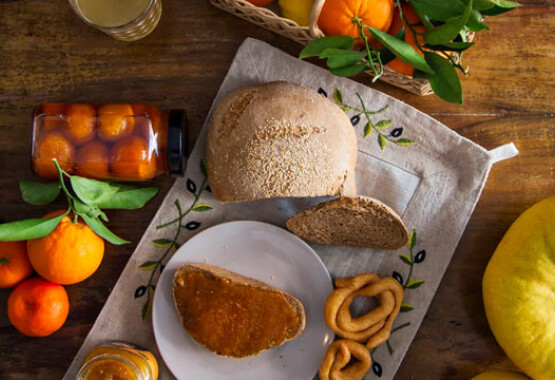 Ξύλινο τραπέζι με ένα καλάθι με εσπεριδοειδή, ψωμί χωριάτικο, πιάτο με ψωμί και μαρμελάδα, χυμό, μαρμελάδα και κουλούρια.