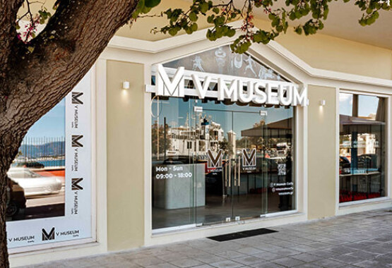 Glass museum facade with the museum's name, framed by a light-colored wall. Reflecting low buildings and a clear sky.