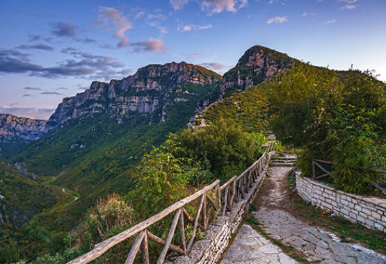 Paved path with a wooden fence and a view of the canyon. Green steep slopes and high cliffs. Clear sky with few clouds in the evening.