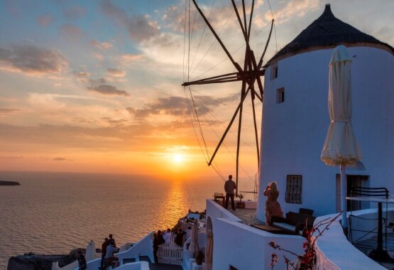 Ηλιοβασίλεμα στην Οία της Σαντορίνης, με παραδοσιακό λευκό ανεμόμυλο να δεσπόζει πάνω από το Αιγαίο Πέλαγος. Άνθρωποι απολαμβάνουν τη θέα από τις βεράντες, ενώ ο ουρανός γεμίζει με ζεστές αποχρώσεις του πορτοκαλί και του ροζ.