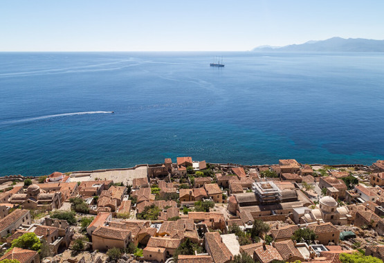 Aerial view of the historic coastal town of Monemvasia, Greece, with terracotta rooftops and the Mediterranean Sea in the background, featuring a distant oil rig.
