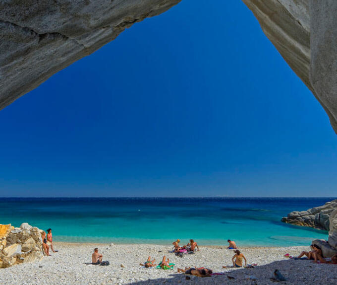 Παραλία με άσπρες πέτρες και γαλαζοπράσινα νερά. Άνθρωποι με μαγιώ στην παραλία. Βράχια τριγύρω σαν σπηλιά.