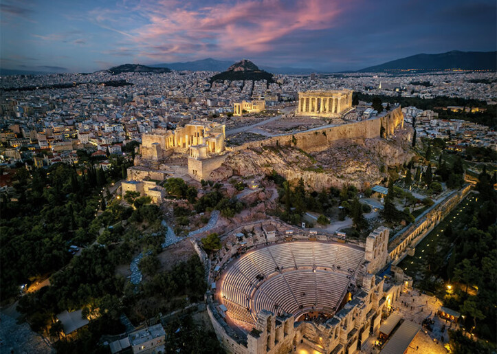 In greek: Aerial view of Athens at dusk, featuring the well-lit Acropolis and Parthenon on a hill, with the expansive cityscape in the background and Lycabettus Hill in the distance.