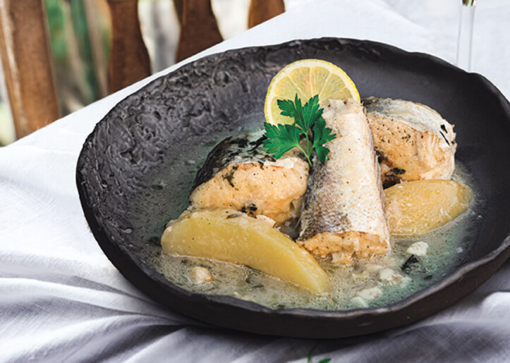 A plate of fish with lemon slices and herbs, accompanied by a glass of white wine, placed on a table with a striped cloth next to a wooden chair.