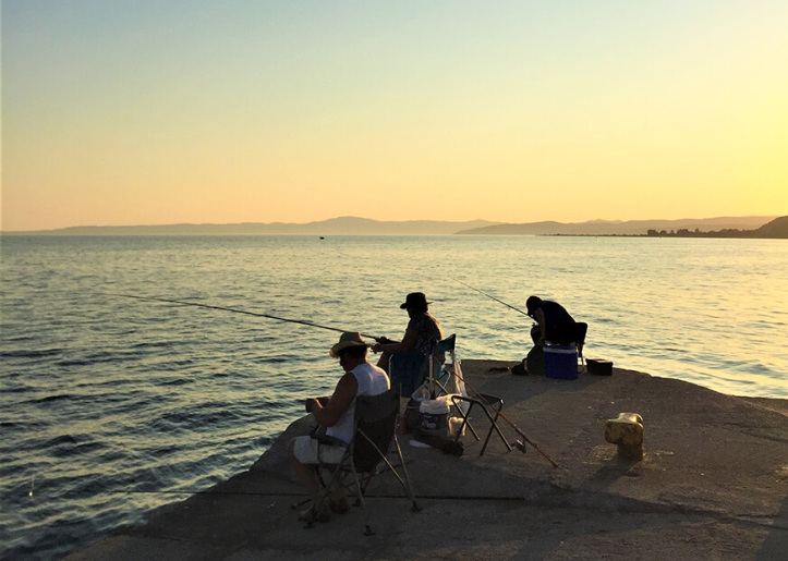 Three people fishing from a concrete pier at sunset with a calm sea and distant hills in the background.