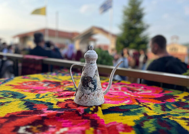 A traditional metal coffee pot sits on a brightly patterned tablecloth at an outdoor setting with blurred people and Greek flags in the background.