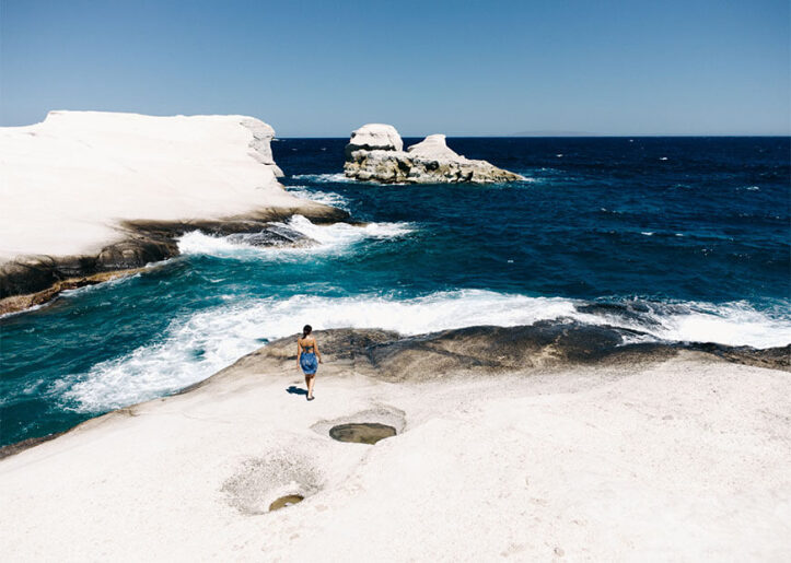 Person standing on the rocky shore of Sarakiniko Beach in Milos, Greece, looking at the bright blue sea and uniquely eroded white limestone formations.