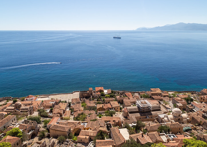 Aerial view of the historic coastal town of Monemvasia, Greece, with terracotta rooftops and the Mediterranean Sea in the background, featuring a distant oil rig.
