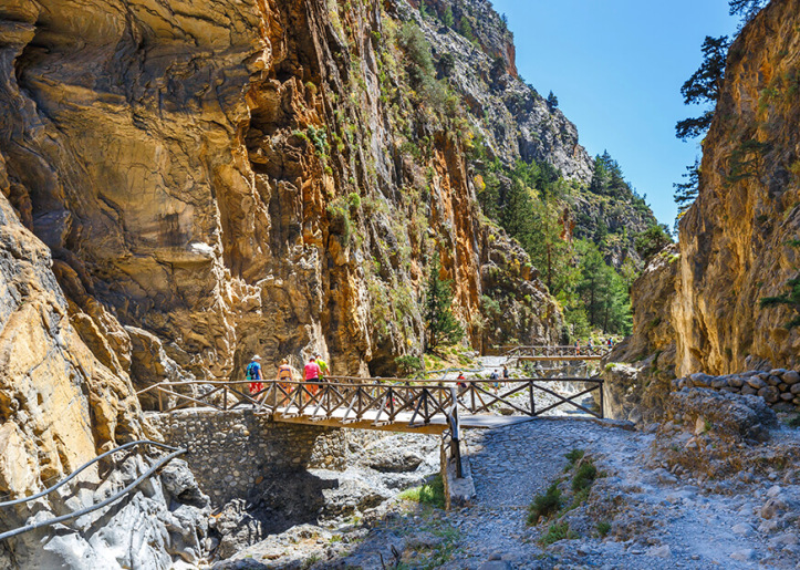 Scenic view of Samaria Gorge in Crete, featuring a wooden bridge crossing a rocky riverbed with visitors walking along the path, surrounded by steep cliff walls and lush greenery.