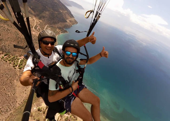 Two people tandem paragliding over mountainous terrain, both smiling and one giving a thumbs-up, under clear blue skies.