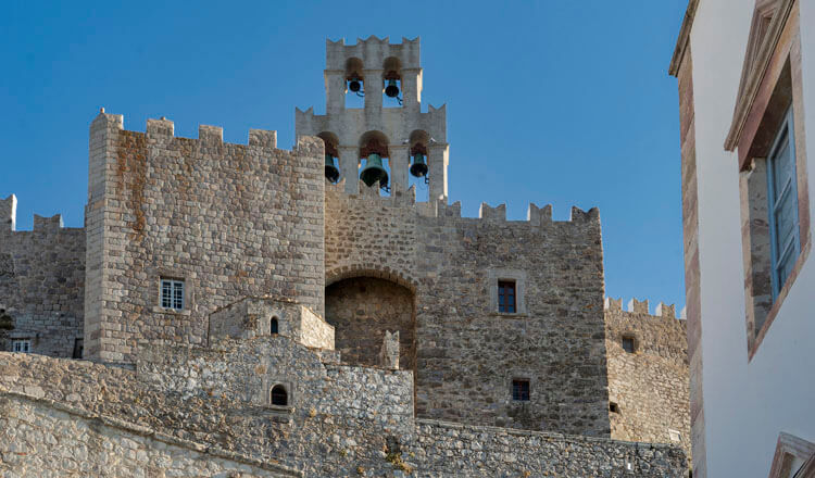 Exterior stone castle walls with small windows and a large bell tower. In front a small stone church. 