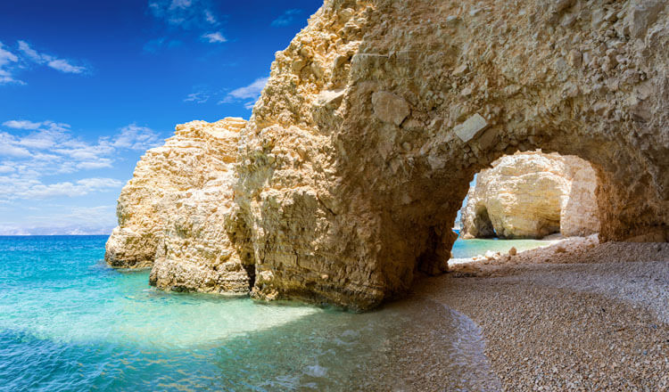 Rock that forms an arch over a beach with small stones and turquoise waters. In the background other rocks.