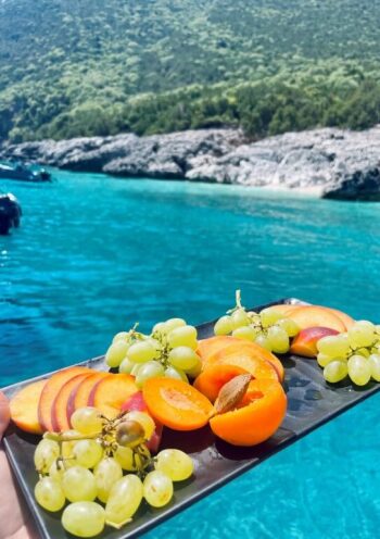 hand holding a platter of fresh cut fruits with turquoise blue sea and rocks in the background