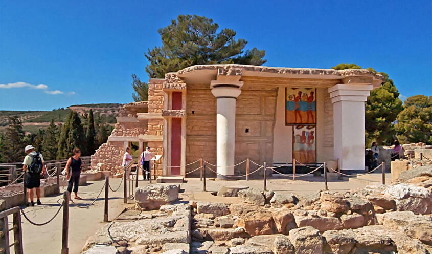 Tourists visit the partially reconstructed ruins of the Knossos Palace in Crete, Greece, under a clear blue sky.