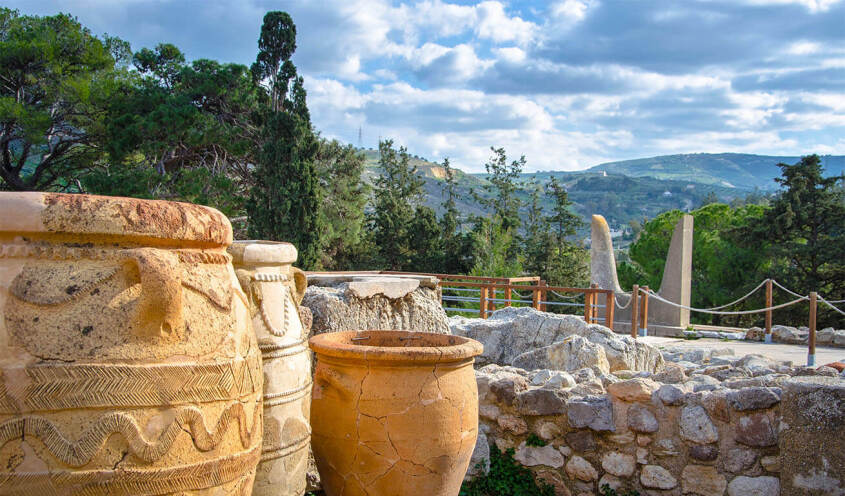 Ancient pottery jars are displayed outdoors amid a scenic, mountainous landscape with trees and cloudy skies.