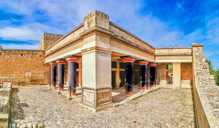 Exterior view of an ancient building with dark colored columns and light colored surfaces. Paved enclosure and stone walls.