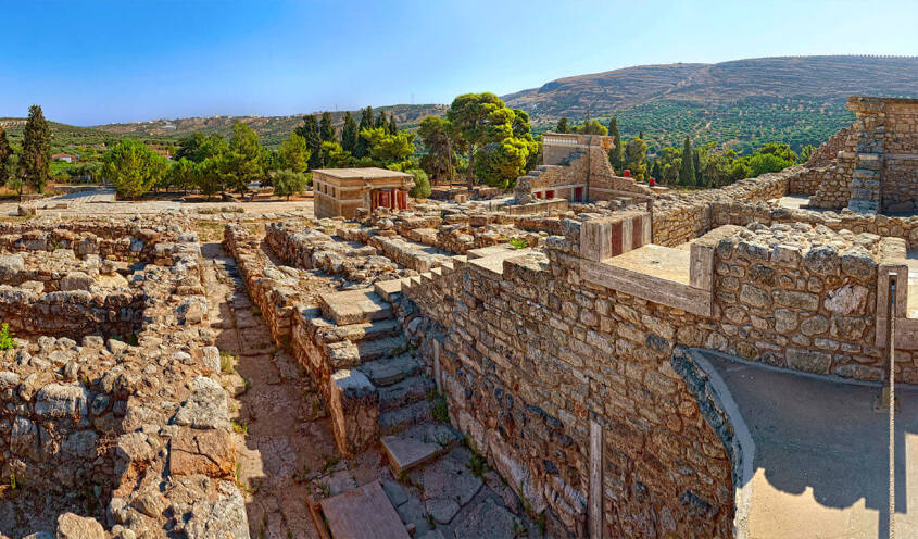 Panoramic view of the ancient ruins of Knossos Palace, with stone structures and surrounding lush greenery under a clear blue sky.