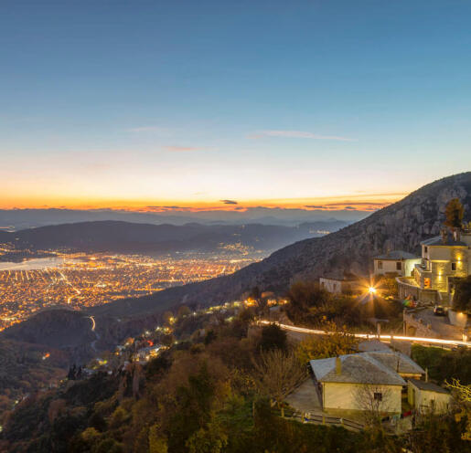 View from above of a seaside town at sunset with lights on. In the foreground traditional houses on the mountain slope. 