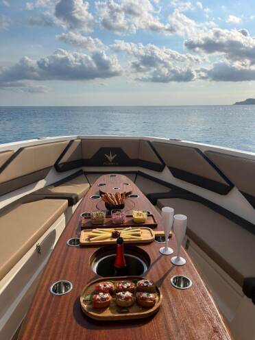 Elegant dining setup on the bow of a yacht with a charcuterie board, cupcakes, and champagne flutes; ocean horizon in the background under a clear blue sky.
