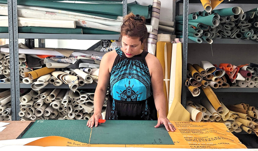 A person at a cutting table measuring fabric, surrounded by numerous rolls of material stored vertically in a workshop