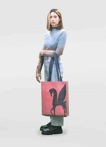 Person standing with a tote bag featuring a black sequined seahorse design, wearing a light blue top, gray trousers, and black platform shoes